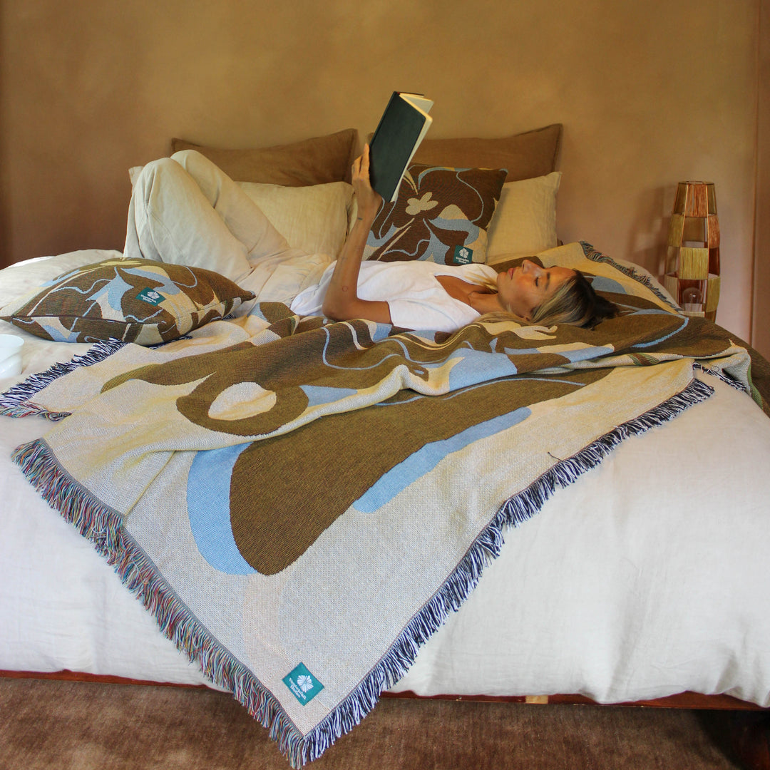 Person lying on a bed with a patterned blanket, reading a book, in a room with a warm-toned wall and pendant light.